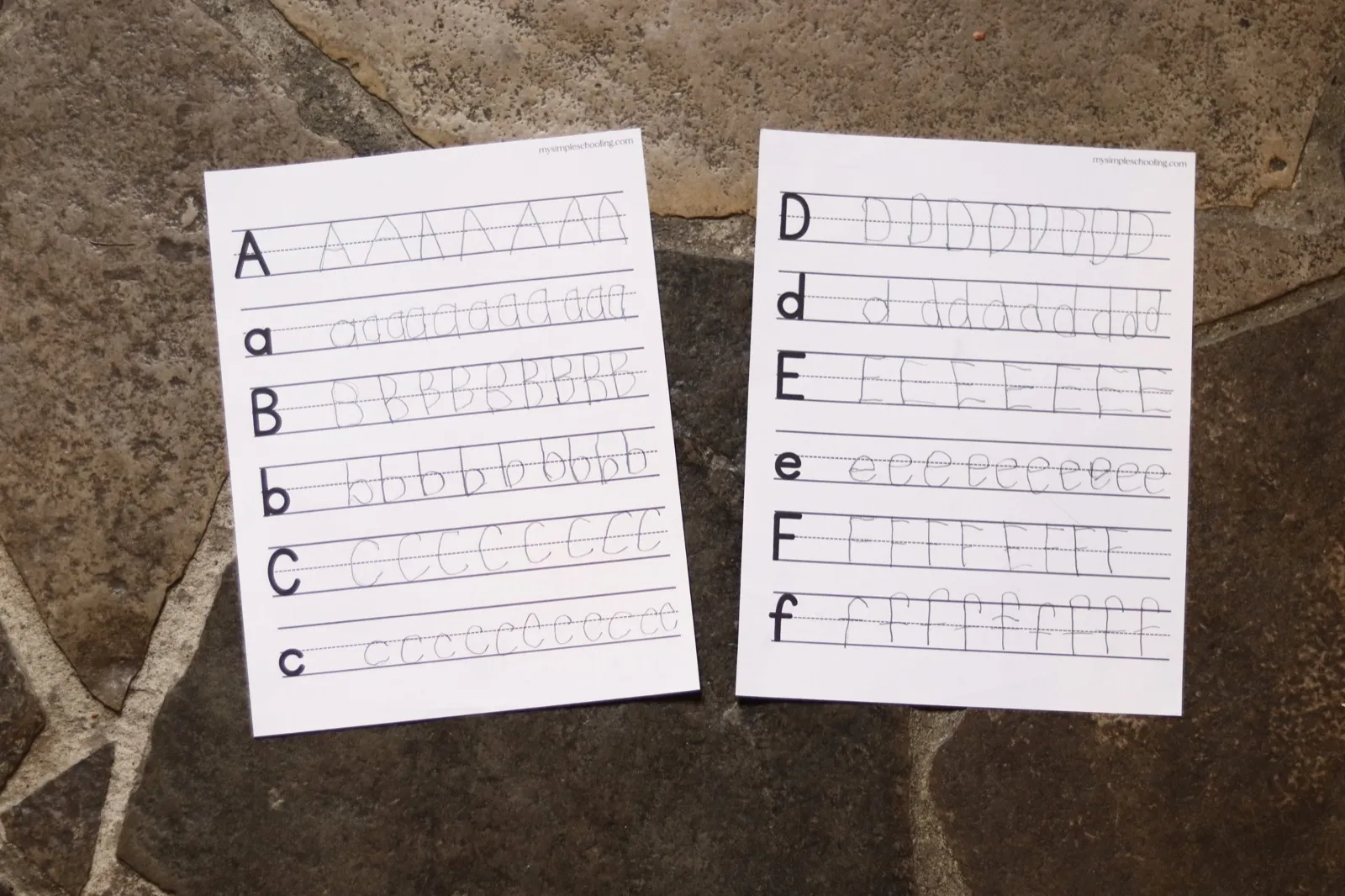 Two completed letter practice sheets filled in by a child, photographed in daylight on a stone floor