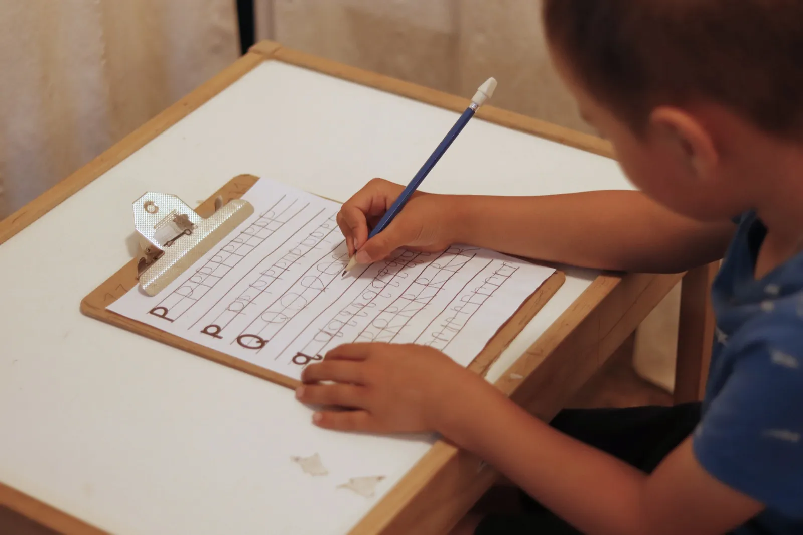 A young child at a small desk, writing letters on a clipboard sheet by himself